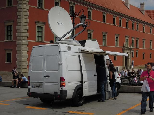 A white utility van with a satellite dish parked in front of a red brick building.